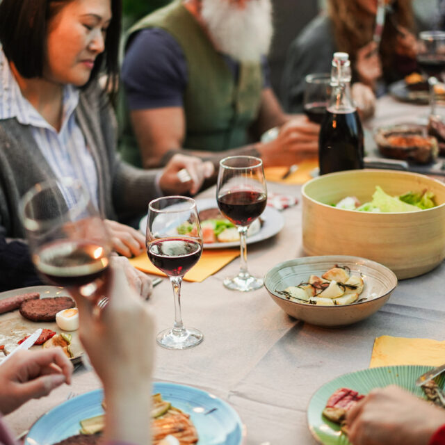가족과 친구들이 야외 바비큐 파티에서 레드 와인을 마시며 식사를 즐기는 모습 - 다양한 연령대의 사람들이 바비큐 저녁 식사를 즐기는 모습 - 여름철 라이프스타일과 즐거움의 콘셉트 - 사진의 주요 초점은 아래쪽 와인잔에 맞춰져 있습니다